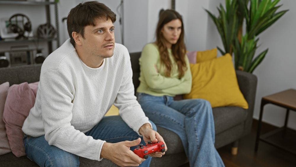 A focused man plays video games as an upset woman watches, symbolizing relationship issues in a living room setting.