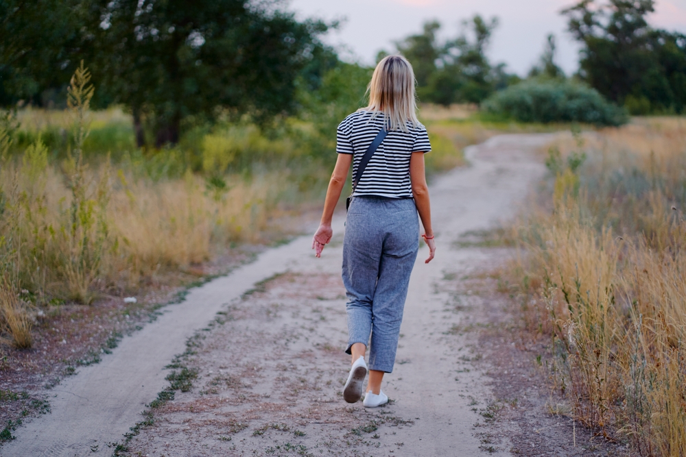 A woman walks on a nature path in a rural area, surrounded by grass and trees. This image evokes feelings of solitude, calm, and reflection in a serene natural setting.