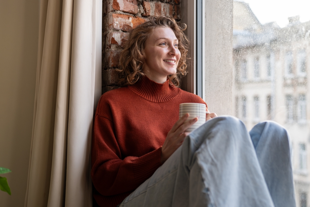 Happy woman enjoying cozy moment with hot drink by window. Smiling female feeling relaxed, takes break from daily tasks with cup of coffee. Mindfulness, self-care, simple pleasures in everyday life. 