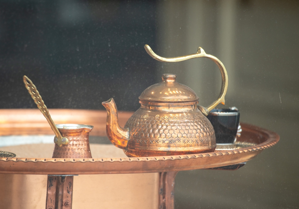 A copper tea kettle sits on a tray next to a small metal spoon. The tea kettle is decorated with a handle and a lid. The tray is made of metal and has a design on it