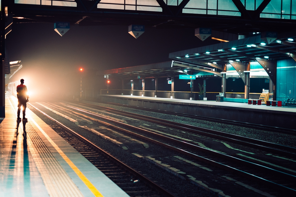 Silhouetted figure of a railway worker standing on a train platform at night as a train approaches with headlights beaming through the fog.