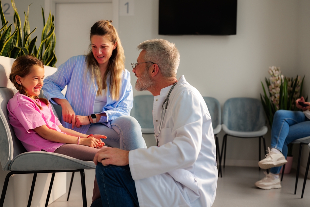 Senior male doctor talking to a young girl and her mother in the waiting room of a modern medical clinic, reassuring them before a consultation