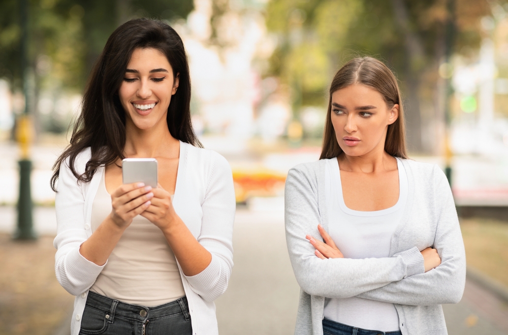 Jealous Girl Looking At Friend's Phone Walking Together Outdoors