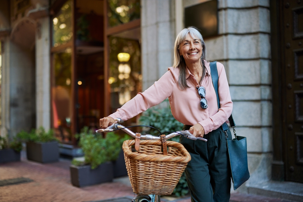 Happy senior woman in casual smiling while holding her bicycle. Older lady walking outside with her bike on an urban scene, copy space. Mature woman enjoying a peaceful walk with cycle in town.