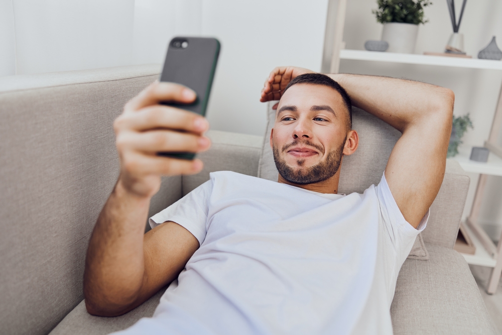 Relaxed man using smartphone at home, enjoying his leisure time. The cozy atmosphere highlights comfort and modern lifestyle. Great for social media themes.