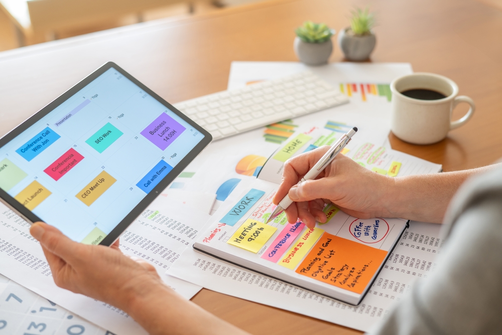 Person balancing financial analysis and daily planning, switching between tablet calendar and paper planner to schedule tasks, track budgets, and organize projects at a desk