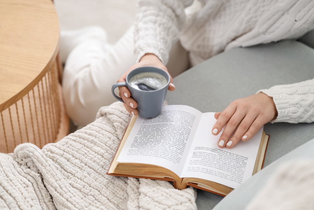 Young woman with cup of hot coffee and book on sofa in living room, closeup