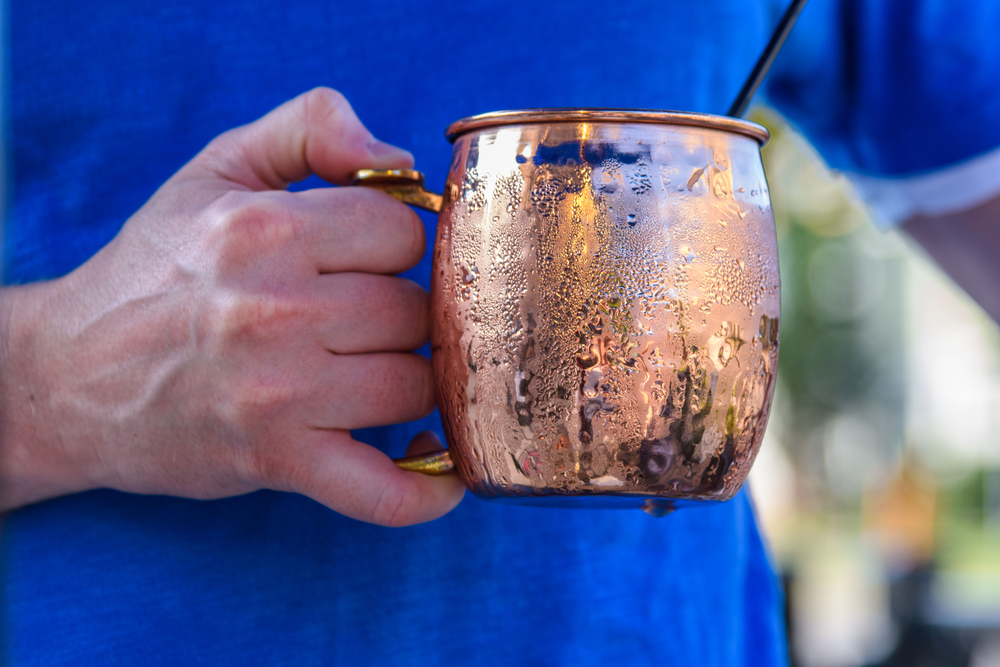 Young man holding a cold refreshing Moscow mule in a bright shiny copper mug outdoor in sunlight - soft focus