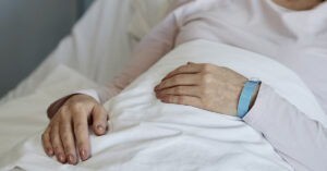 Elderly woman lying in hospital bed, holding blanket with one hand and wearing medical bracelet on wrist. Gentle sunlight illuminating soft bedding