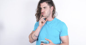 Narcissists Can Be Kind Too. Don’t Mistake It For Goodness - Narcissistic Kindness Isn’t What It Seems Young caucasian man with long hair wearing blue t-shirt over white background thinks deeply about something, uses modern mobile phone, tries to made up good message, keeps index finger near lips
