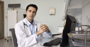 professional male doctor in a white lab coat sitting at a desk and typing on a computer, likely working on electronic medical reports