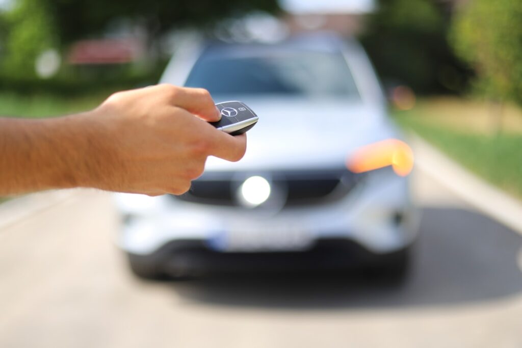 A hand holds a car key fob in the foreground with a silver car blurred in the background on a sunny residential street.