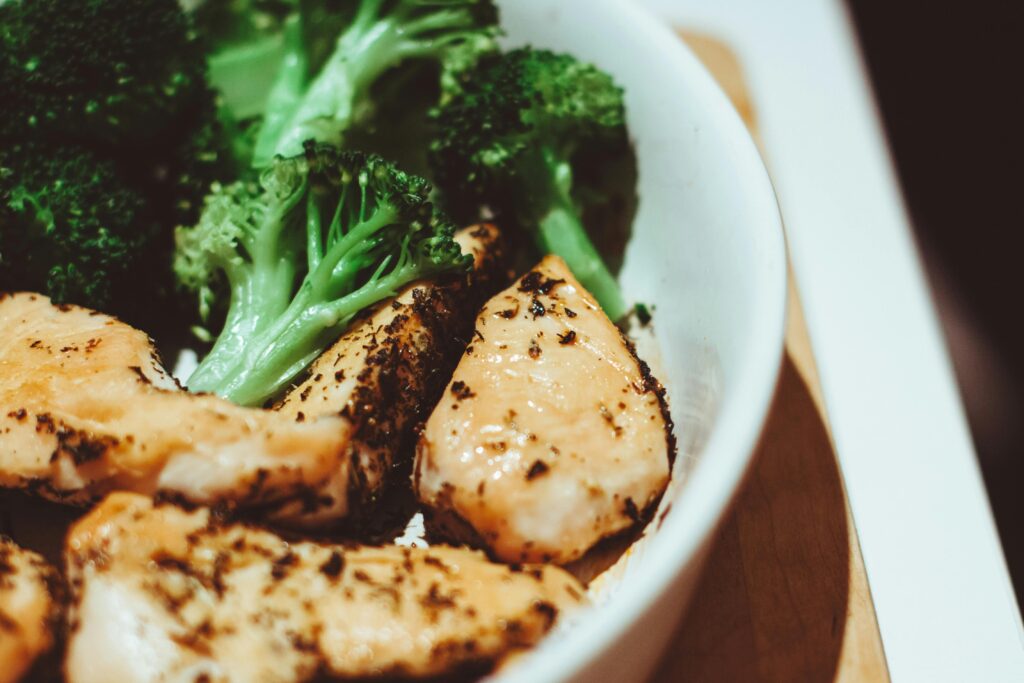 A close-up photo of a white ceramic bowl on a surface containing pieces of seasoned grilled chicken breast alongside fresh bright-green broccoli florets, with visible herbs and spices on the chicken.