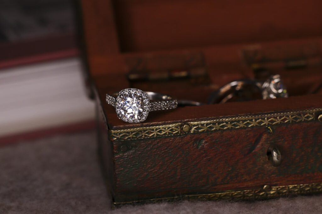 A diamond engagement ring with a halo setting rests inside an open antique jewelry box lined with worn red velvet.