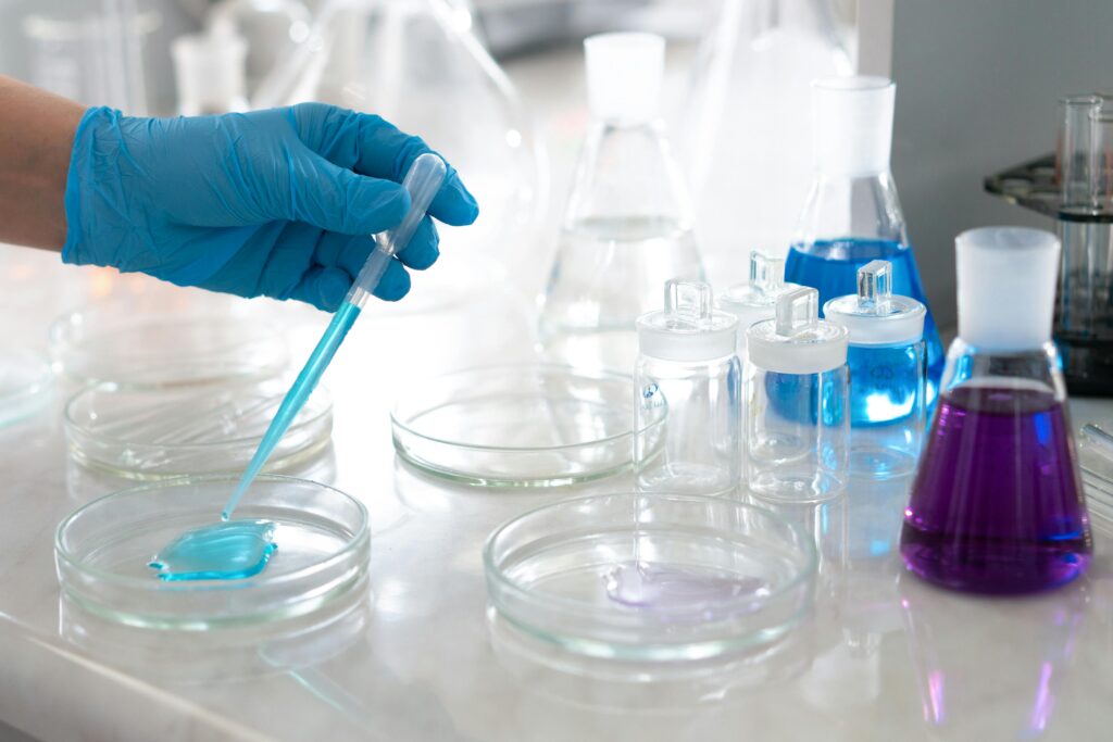 A gloved hand uses a pipette to dispense blue liquid into a petri dish inside a laboratory. Glass beakers and flasks containing blue and purple solutions sit on the white countertop in the background, suggesting pharmaceutical quality testing or chemical analysis.
