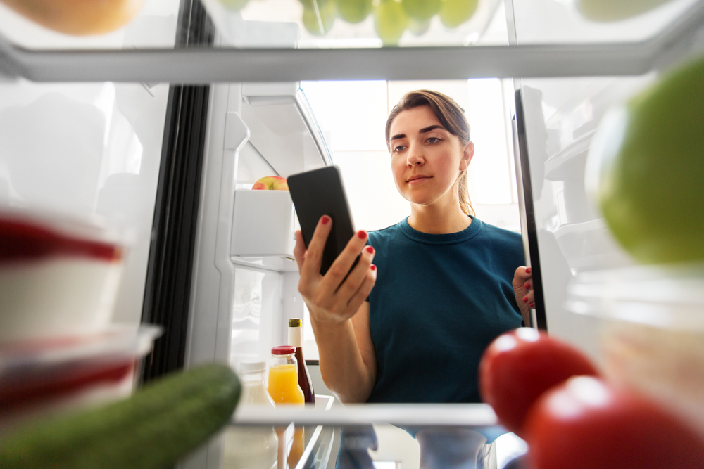 healthy eating and diet concept - woman at fridge with smartphone making list of necessary food at home kitchen
