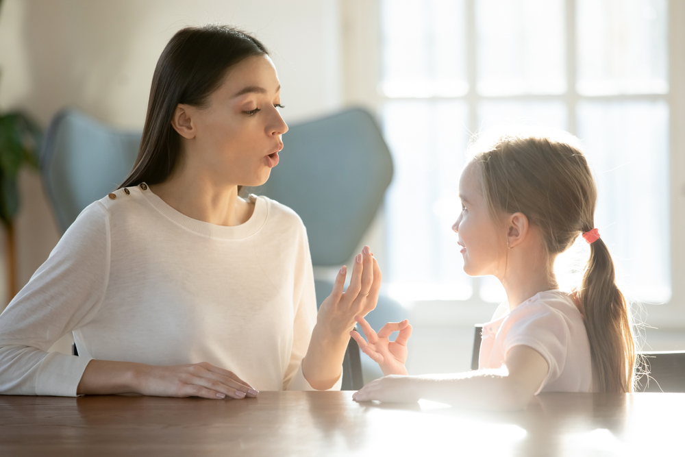 Caring young Caucasian mom talk speak learn pronouncing sounds with small 7s daughter. Millennial female teacher or tutor make hand gesture practice nonverbal sign language with little girl child.