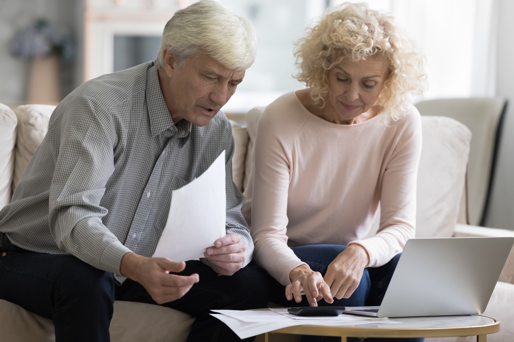 Focused retired married couple checking paper bills, invoices, paying medical insurance fees, doing domestic paperwork, counting income, expenses, reviewing documents