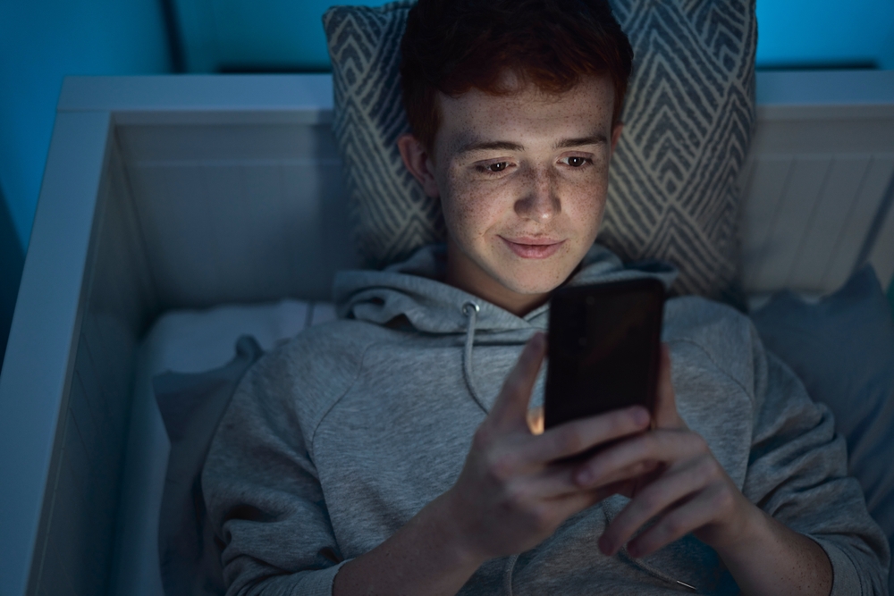 Cheerful caucasian teenage boy using mobile phone while lying in bed at night in his room 