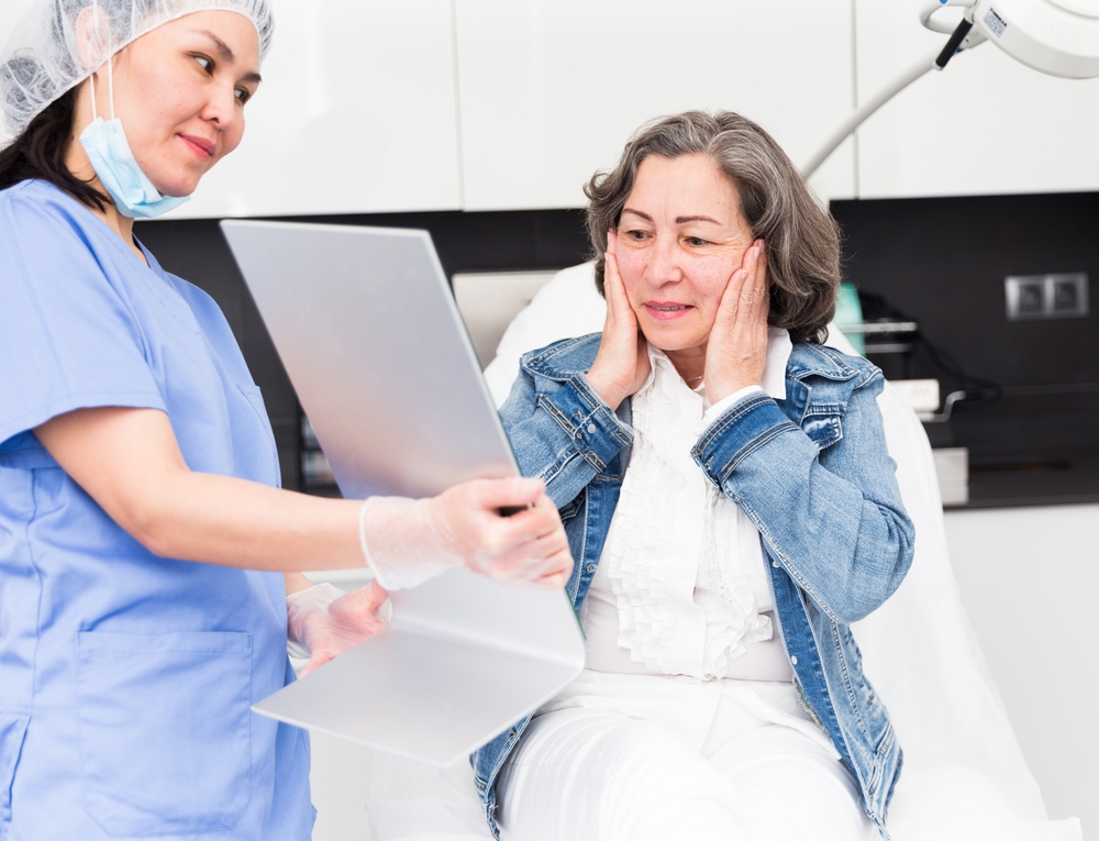 Elderly female client sitting in medical cosmetology office and looking at mirror satisfied with results of facial skin care procedure.