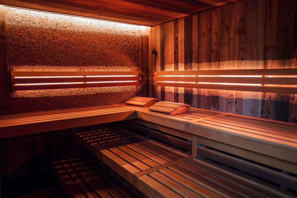 Interior of wooden sauna with light colored walls and benches, multiple levels of seating, infrared heaters emitting soft red glow, and cozy atmosphere.