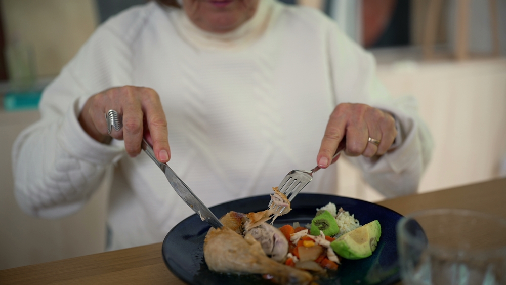Close-up of an older woman cutting food on a plate, enjoying a nutritious meal with avocado, carrots, and chicken, emphasizing healthy eating habits and nutrition for seniors