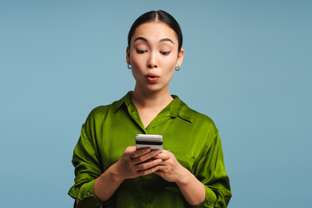 Young woman is looking at her phone with a shocked expression on her face, surprised by what she is reading. Online technology concept