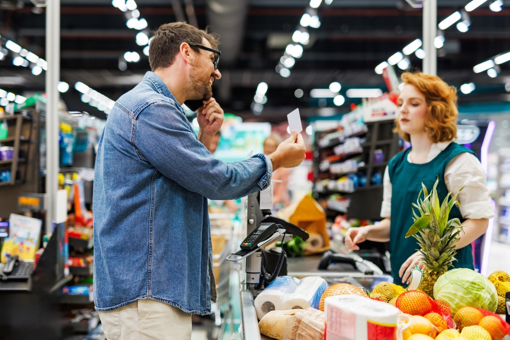 Customer is talking on his cell phone while showing a loyalty card to the cashier at the supermarket checkout, with fresh produce in the foreground