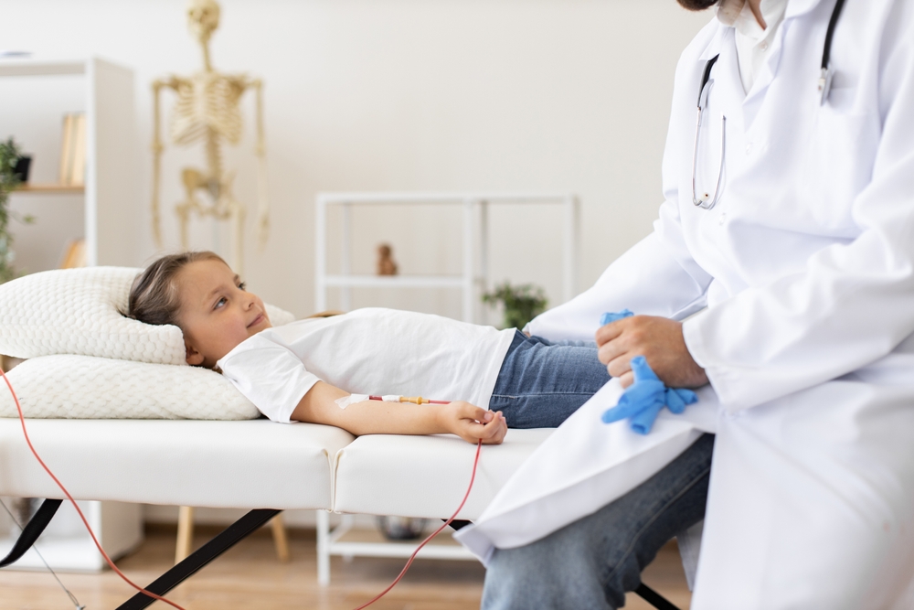 Doctor gives blood transfusion to sick little girl at medical facility. Young girl lying on bed in hospital room receiving treatment from male doctor wearing lab coat.