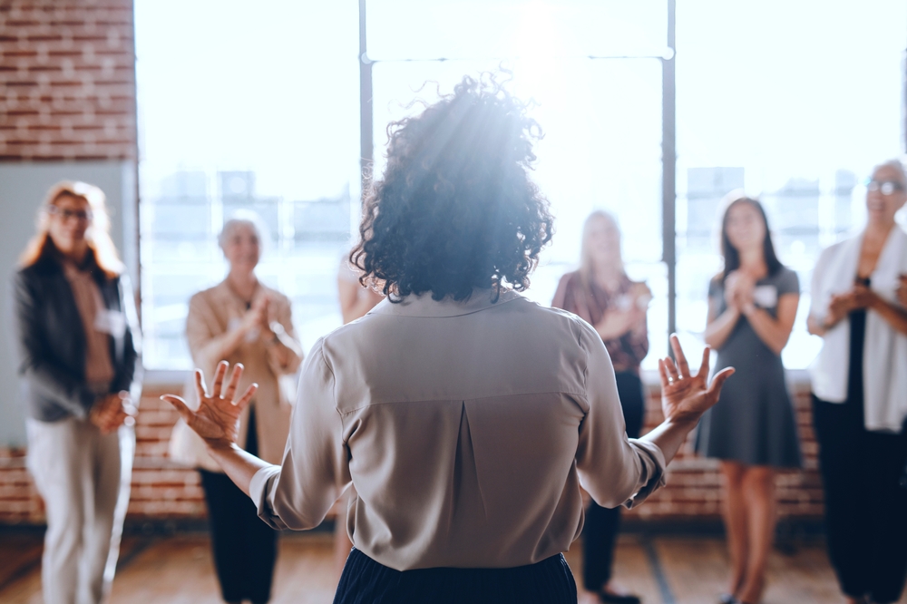 A person stands confidently in front of a group, leading a discussion. The group listens attentively, showing engagement and interest in the presentation. Corporate office team and colleagues.