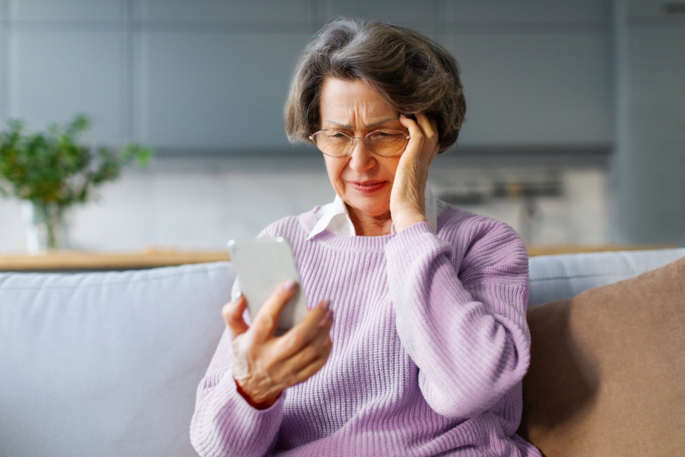 Elderly woman expressing confusion while looking at her smartphone, possibly dealing with technology issues or reading bad news