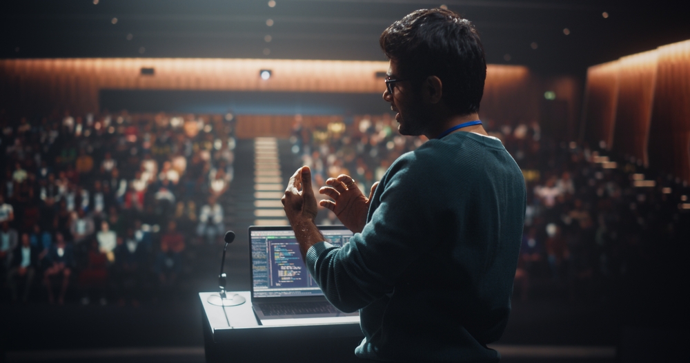 Portrait of a Young Speaker Standing on a Conference Stage. Technology Startup Founder Using a Laptop Computer, Speaking About Software Integration at a Business Meeting with Professional Audience