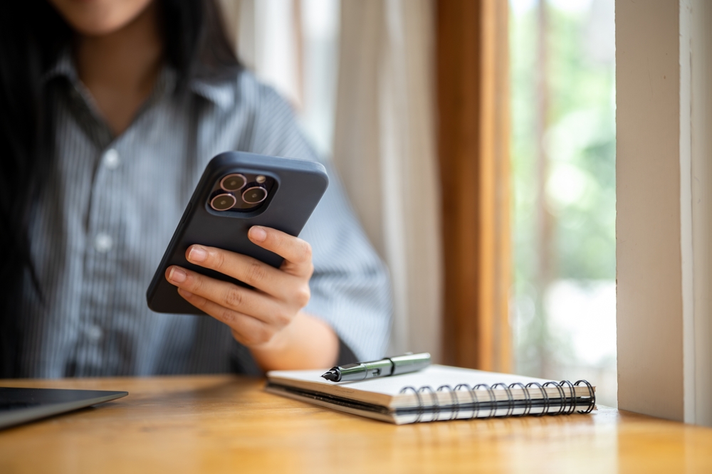 Close up of woman's hand holding or touching on smartphone with pen atop notebook and laptop at cafe wooden table. Working or Study. Chilling Outside.