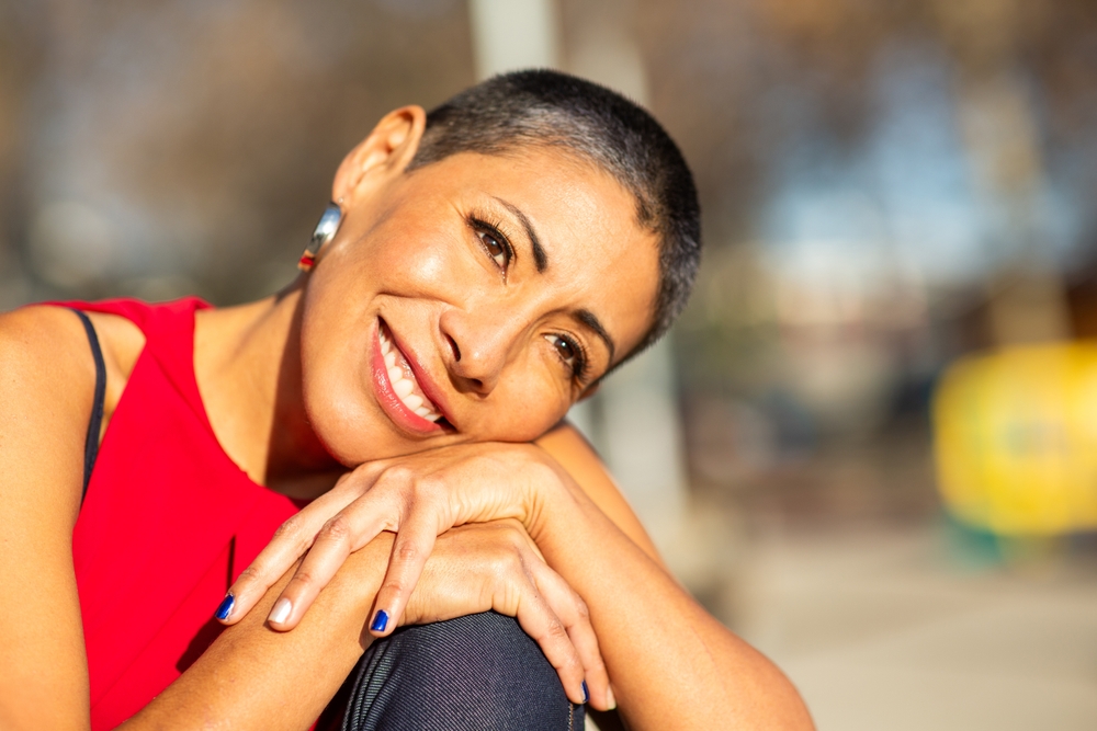 Close-up of a smiling woman with a shaved hairstyle and bright red blouse leaning her face on her hands while looking into the distance