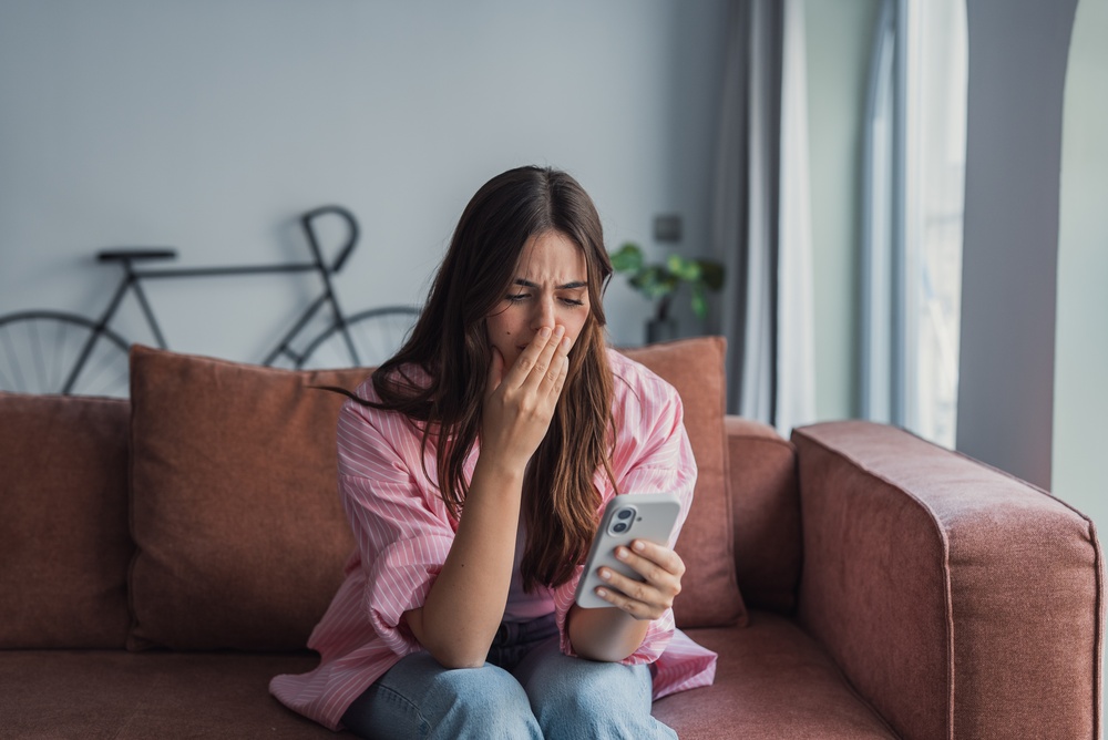 A frustrated millennial Latin woman sits on the sofa reading sudden bad news on her phone, covering her face with her hand in despair after receiving a threatening message or upsetting email