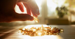 A man's hand reaching for yellow transparent vitamin D and Omega-3 fish oil capsules in the sunlight on a bright, sunny table