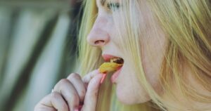 10 Weird Signs That May Indicate You're Sleep-Deprived (Beyond Fatigue) Blond woman biting crunchy potato chip, smiling while tasting salty snack during sunny outdoor moment