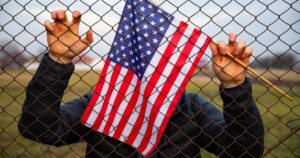 New Crackdown Could Strip “Hundreds” Of Americans Of U.S. Citizenship Man holding American Flag near a barbed wire, USA border. American immigration and United States