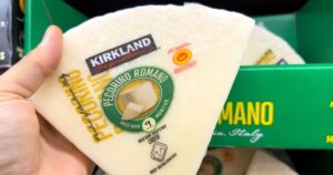 Costco vs Walmart, Which Store Actually Saves You More Money Hand holding large triangular piece of pecorino romano cheese made from sheep milk and aged in sardinia italy at Costco Wholesale retail warehouse