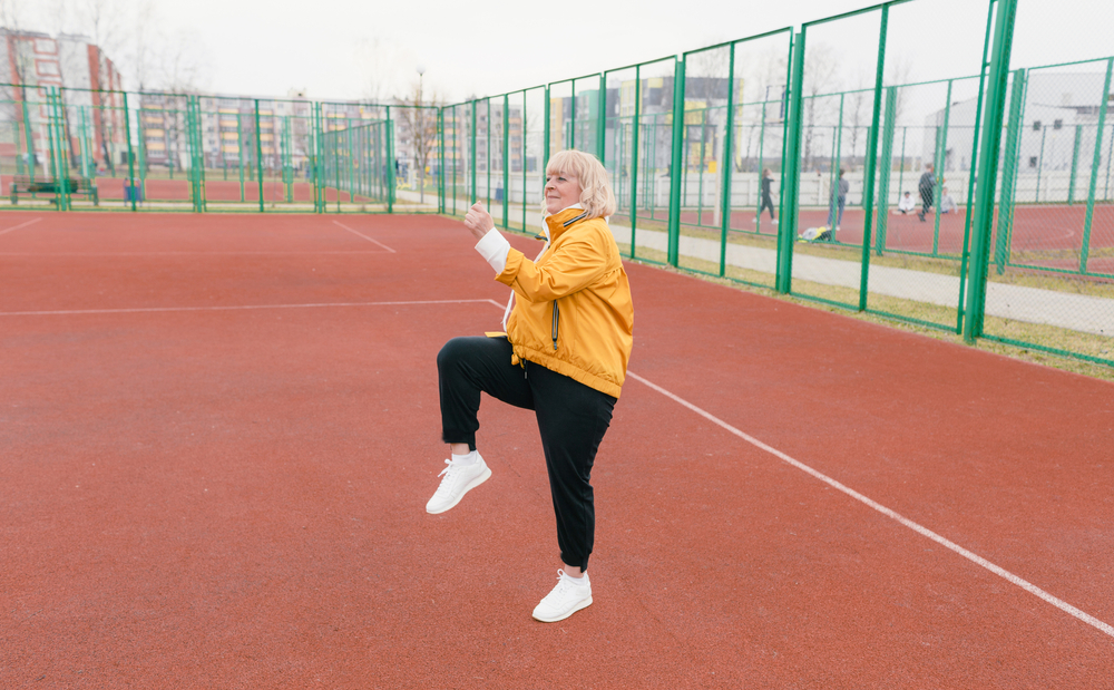 an elderly woman in a yellow jacket is doing sports exercises on a red treadmill. the stadium is a healthy lifestyle. retired people and sports. active old woman