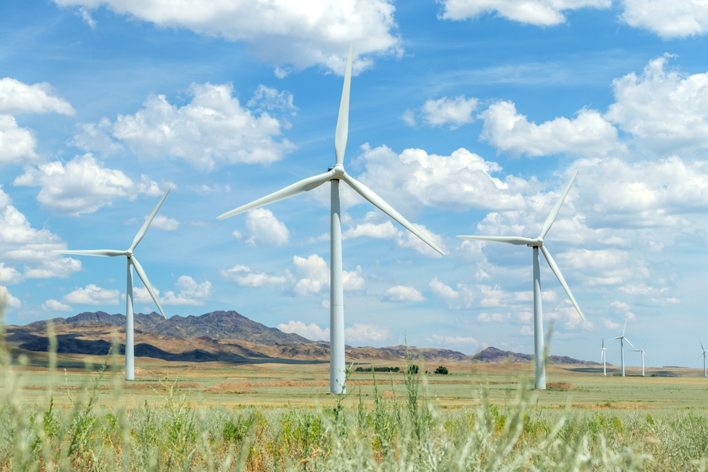 Several tall wind turbines spinning in a remote grassy field surrounded by arid hills and mountains. An excellent image for renewable energy, sustainability, or environmental concepts.
