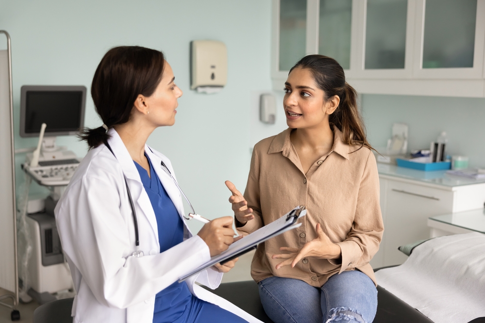 Young 20s Indian patient woman speaking to female practitioner in ultrasound diagnosing exam room, telling about symptoms, healthcare. Doctor writing notes on consultation meeting