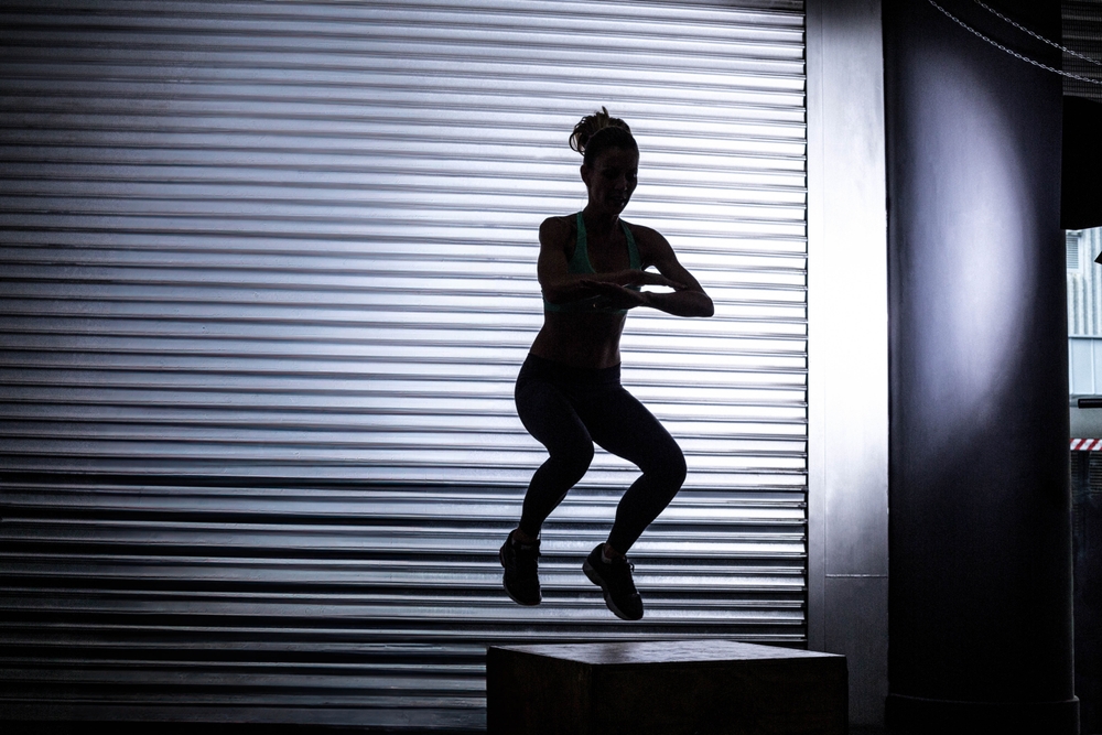 Mid adult female athlete jumping from plyometric box in gym with roll-up shutter door. Dynamic, strength, determination, performance, fitness, workout, industrial
