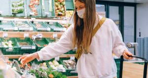 11 Awful Grocery Shopping Habits That Are Costing You More Than You Think Woman wearing a face mask shopping for vegetables in a grocery store during the pandemic.