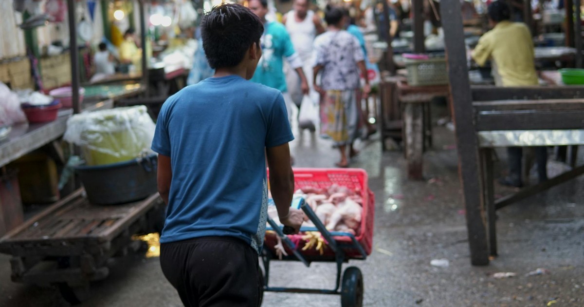 A man pushes a cart filled with goods through a bustling outdoor market, surrounded by shoppers and vendors.