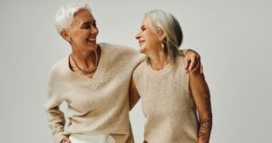 fashionable senior female friends smiling at each other while posing with hands in pockets on grey