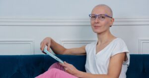 A bald woman struggle of cancer with glasses lounging on a blue velvet sofa while browsing a tablet at home