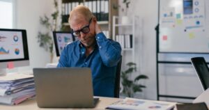Does It Matter What You Do While You Sit? New Dementia Research Says Yes Tired mature entrepreneur experiencing neck pain while sitting at desk in modern office, surrounded by paperwork and computers, highlighting the physical strain of a demanding work environment