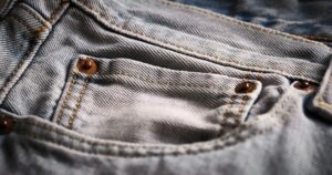 Close-up of a denim jeans front pocket with metal rivets and visible stitching. Fabric texture, seams, worn cotton surface. Durability, casual style, and everyday clothing details