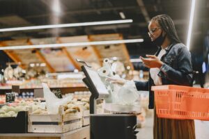 A woman with a mask shops and weighs groceries in a modern supermarket setting.